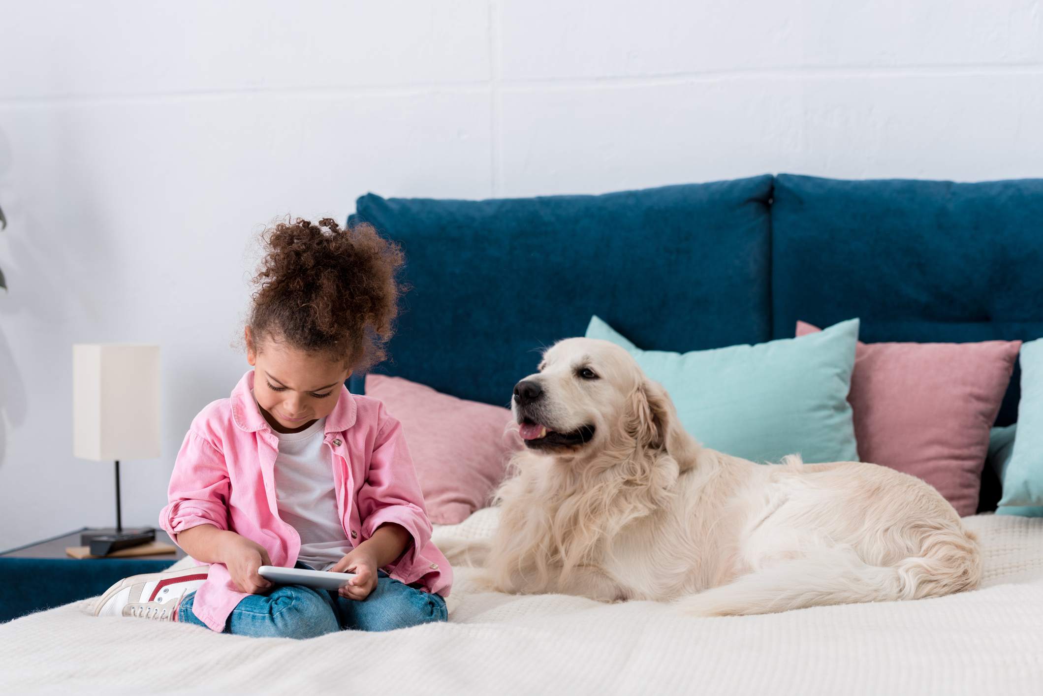 一个面带微笑的非裔美国小孩和金毛猎犬一起坐在床上玩智能手机